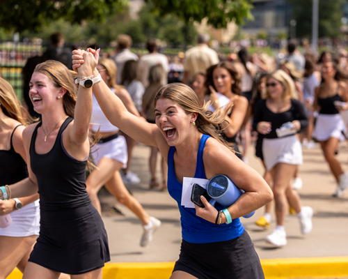 Two women run together outdoors, smiling and cheering with their clasped hands raised in celebration, while a larger group of runners follows behind them.